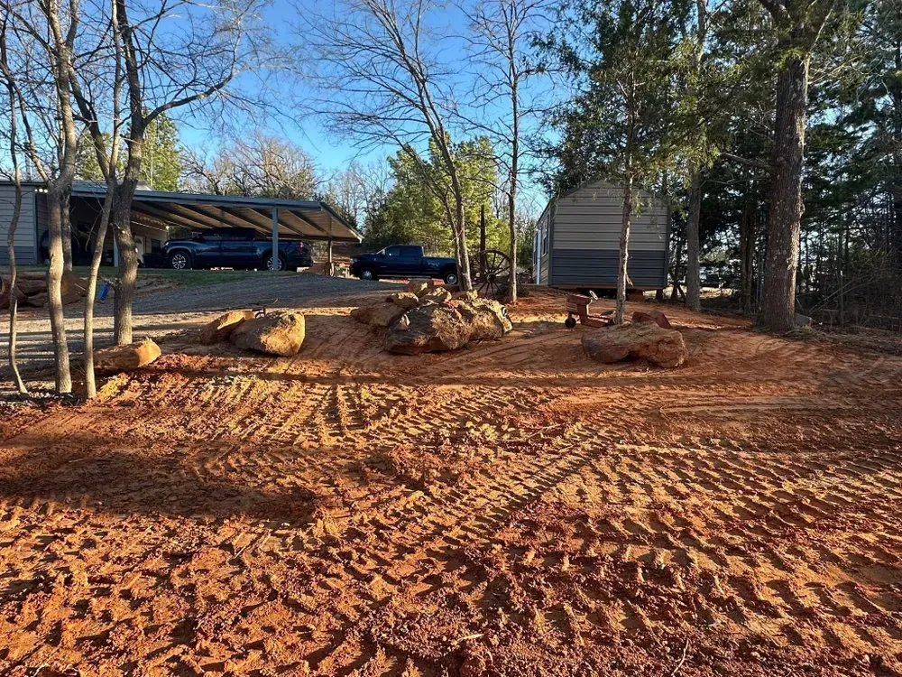 Freshly graded red dirt yard with tree stumps, large rocks, and a house and garage in the background