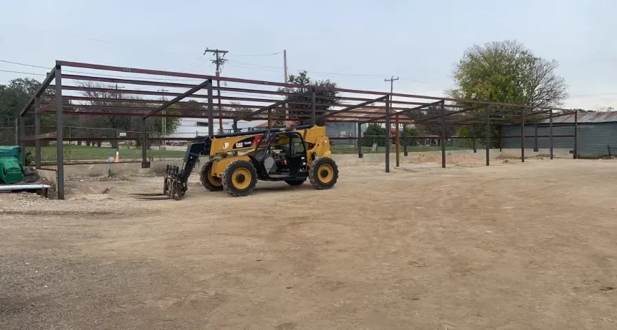 Yellow backhoe loader parked in a dusty yard beside a steel-framed building under construction.