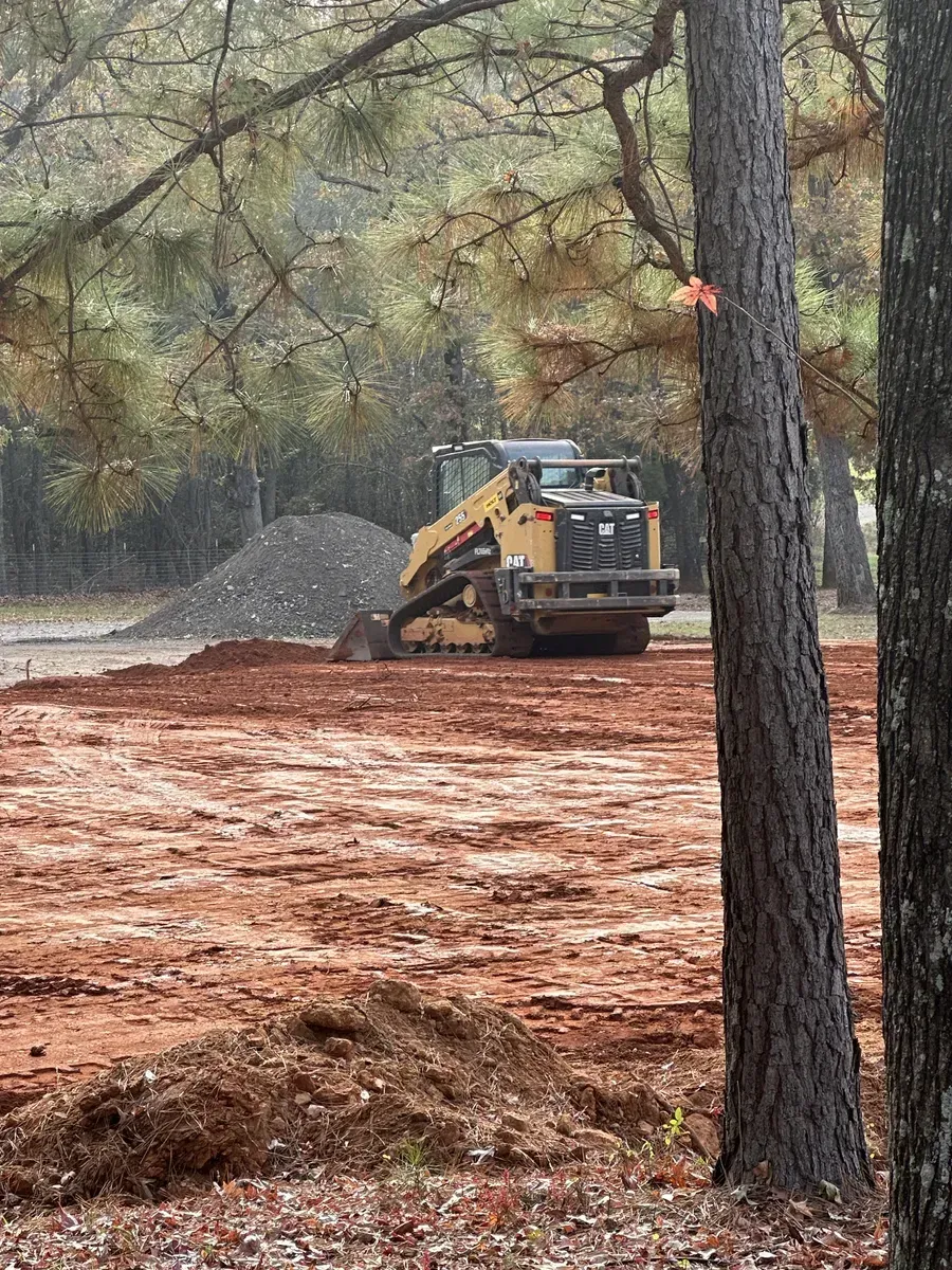 Small construction machine grading red dirt near trees and a gravel pile in a wooded area