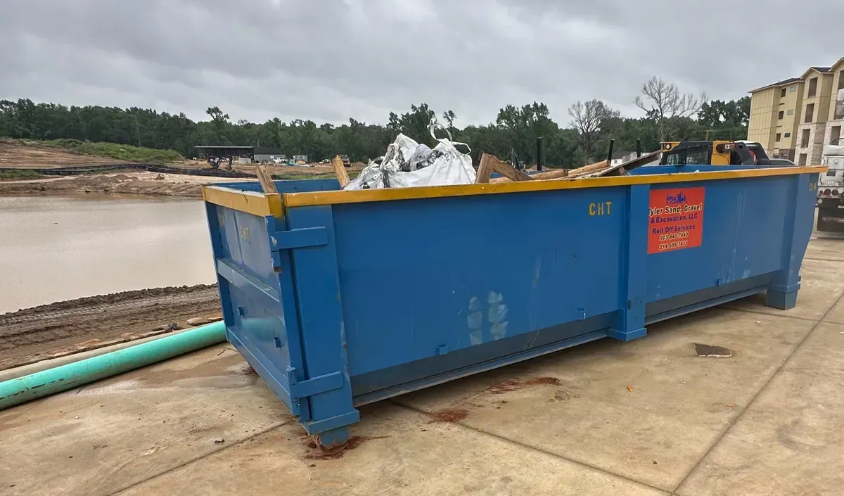 Blue dumpster filled with white debris beside a muddy river on a concrete dock