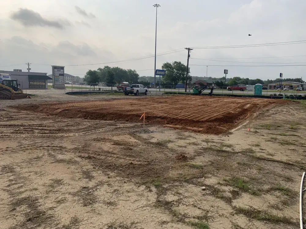 Dirt lot with a freshly graded rectangular area, parked vehicles, and utility poles under a cloudy sky