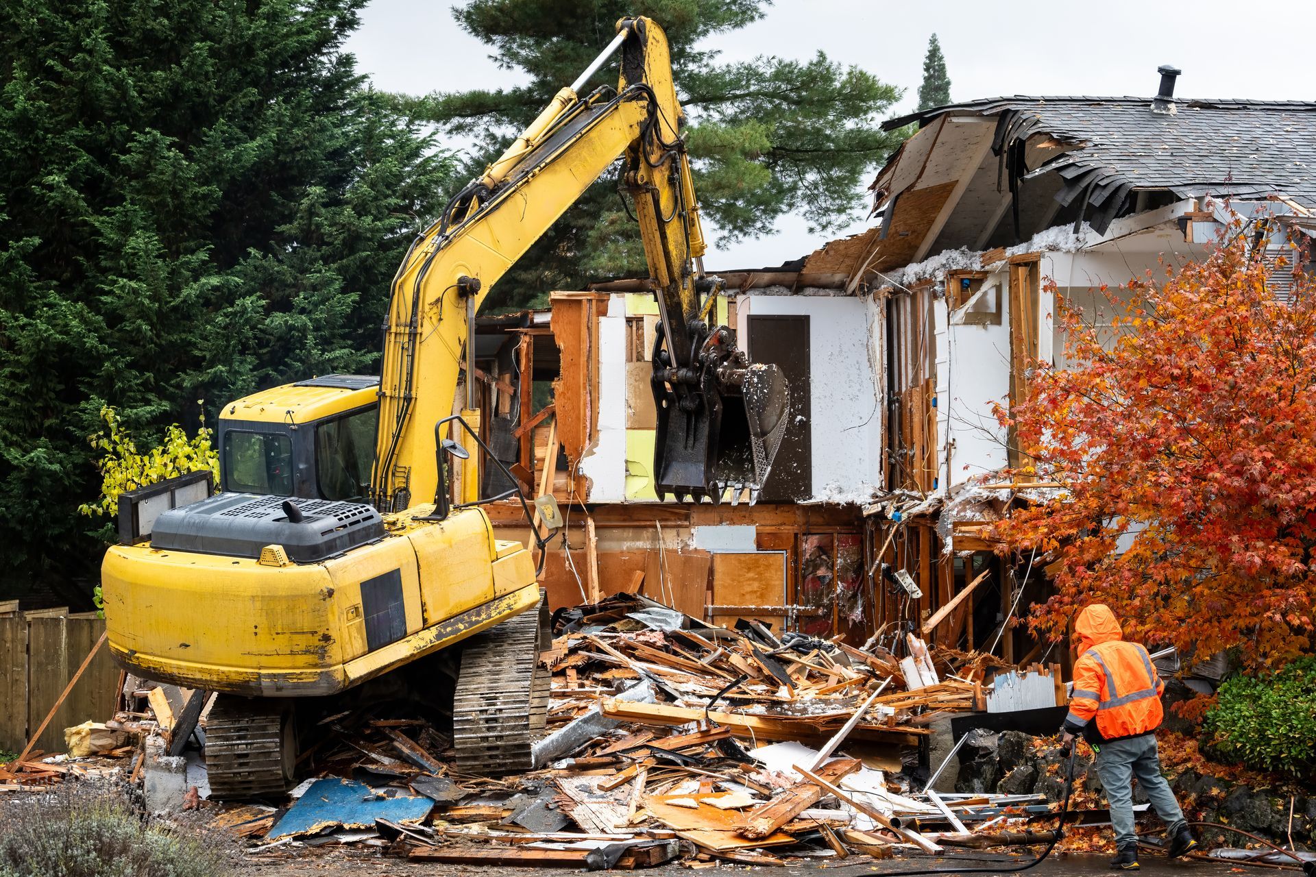 Yellow excavator demolishing a damaged house, with debris and a worker in orange safety gear.