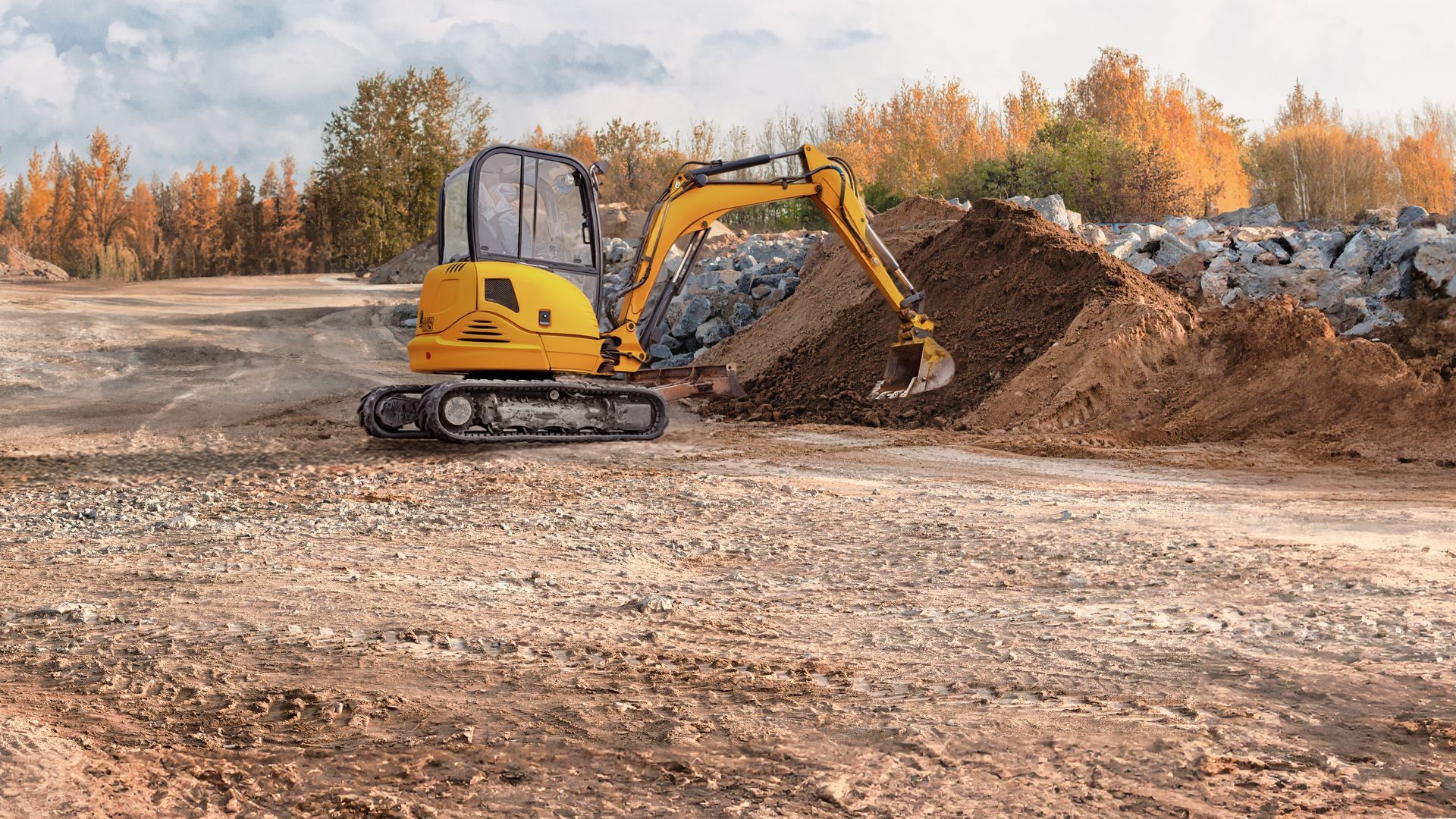 Yellow excavator digging a dirt pile at a construction site with autumn trees in the background