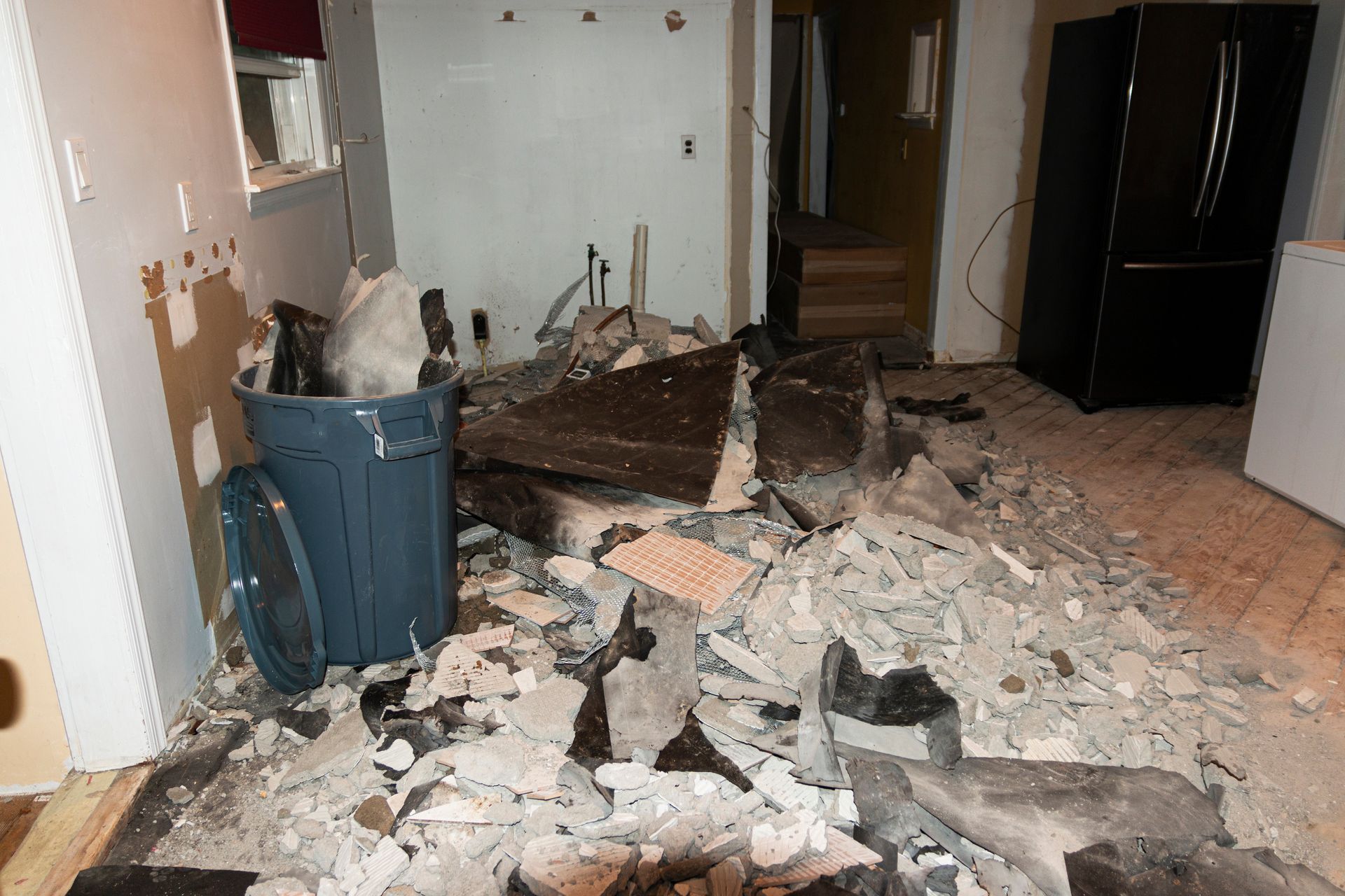 Living room with severe floor damage, rubble, and debris piled near a trash bin.