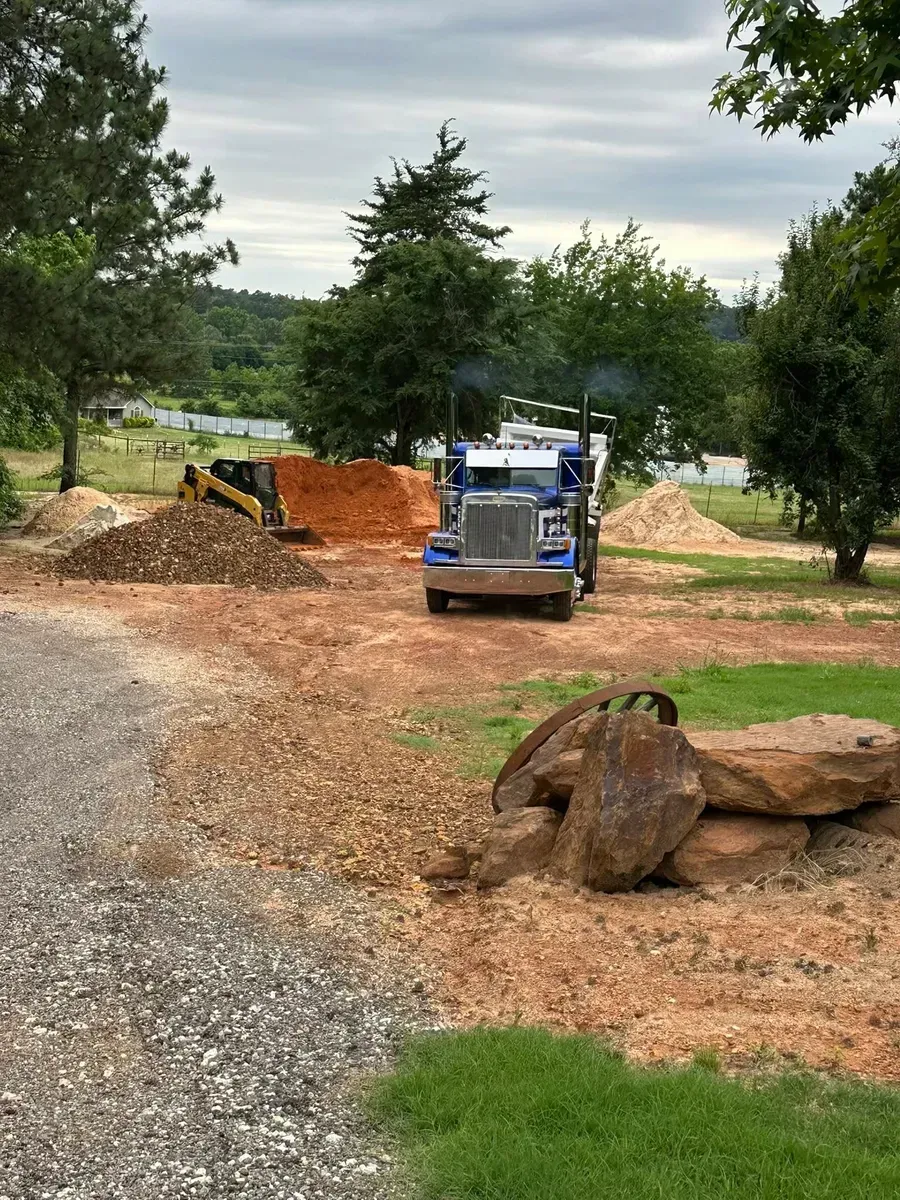 Blue skid steer on gravel path beside dirt piles and rocks in a wooded yard