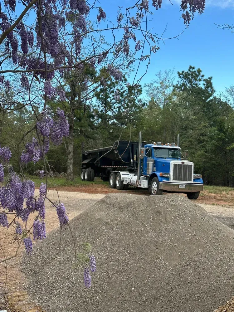 Blue semi-truck hauling gravel on a rural dirt road beside purple flowers and trees
