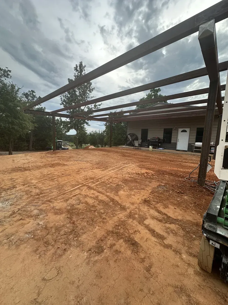 Large dirt lot with steel beams overhead beside a house under construction on a cloudy day