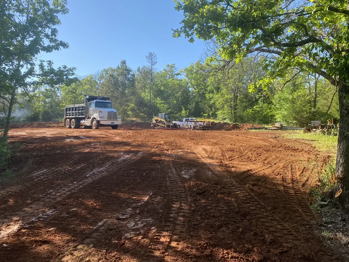 Truck and excavator on a muddy dirt lot surrounded by trees under a clear blue sky
