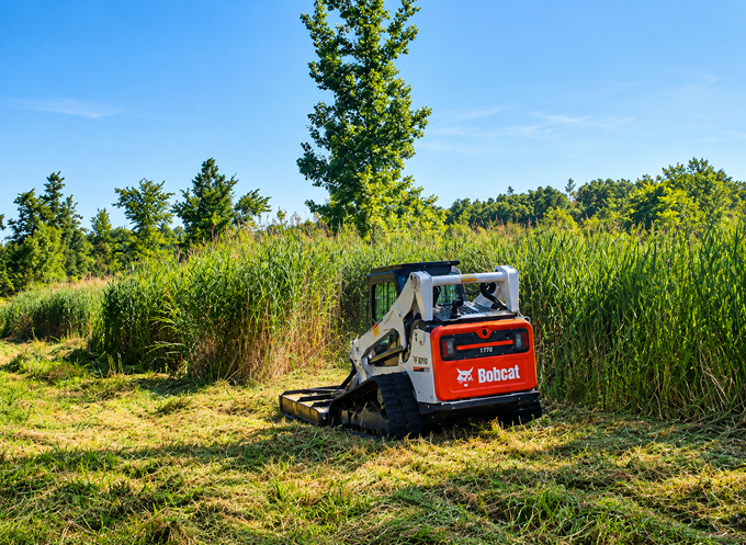 Small red skid-steer mowing tall grass in a sunny field under a blue sky