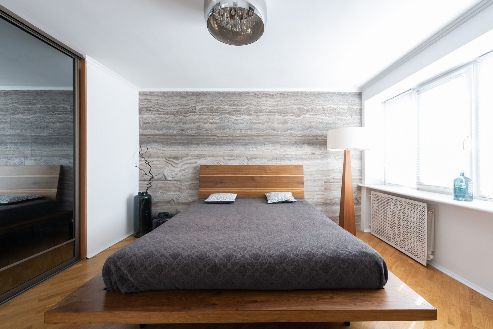 Modern bedroom with a wooden bed, gray blanket, striped accent wall, and large window with radiator below