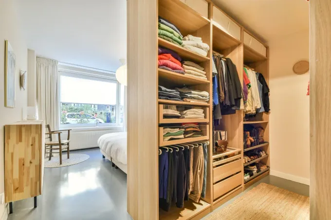 Walk-in closet with open shelving, hanging clothes, and a glimpse into a bright bedroom beyond