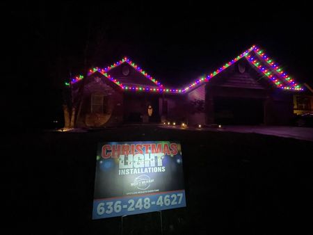 House at night outlined with colorful Christmas lights and a yard sign for lighting services with phone number.