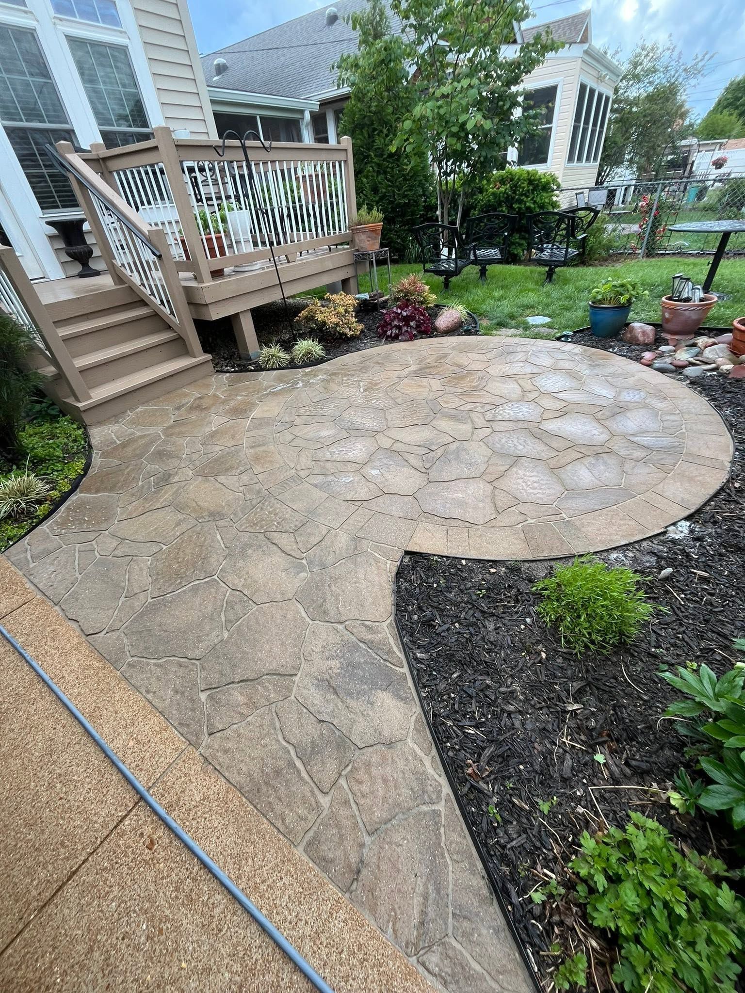 Stone patio and curved walkway beside a wooden deck in a landscaped backyard garden.