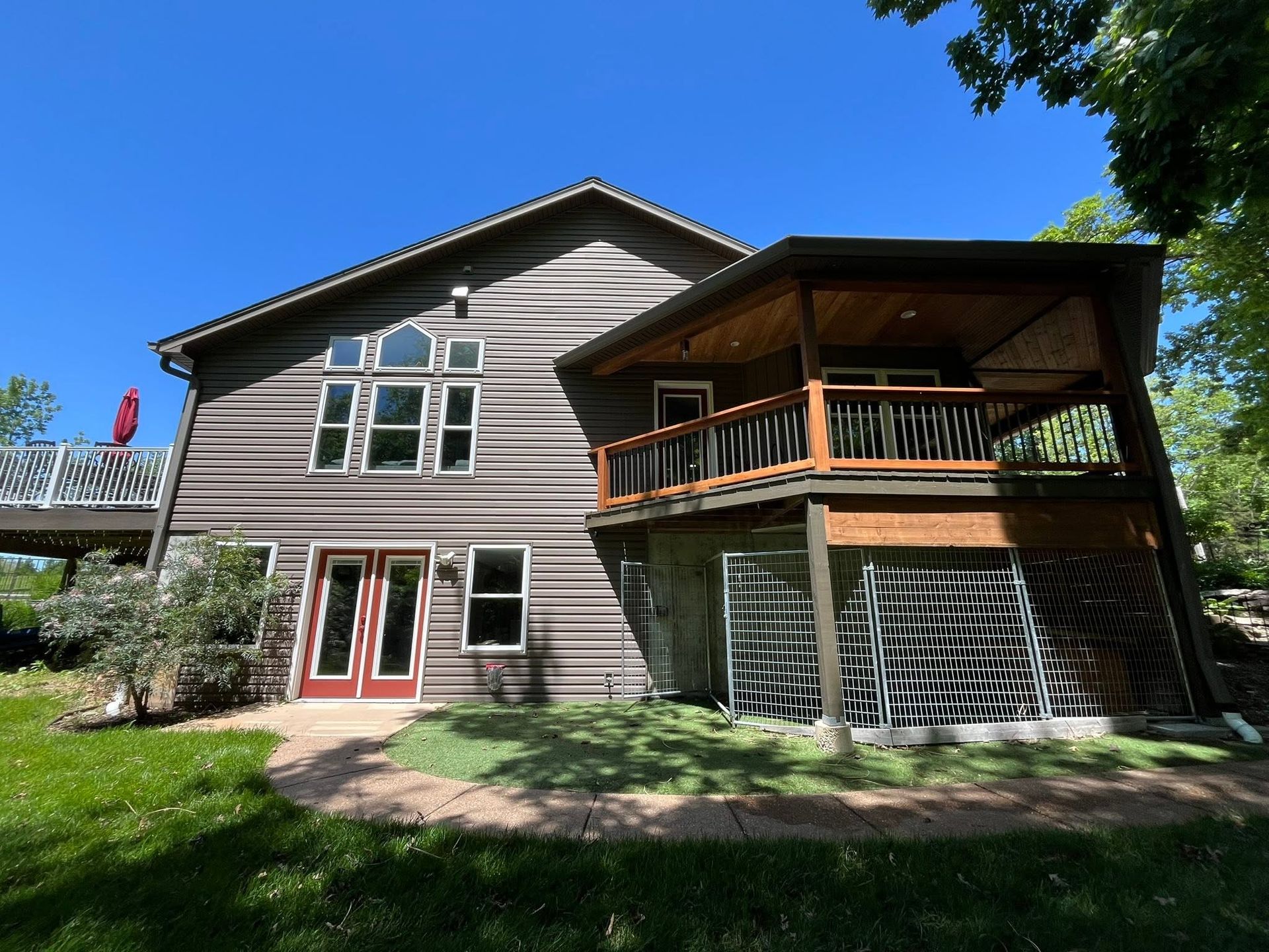 Two-story house with gray siding, red doors, and a wooden balcony under a bright blue sky