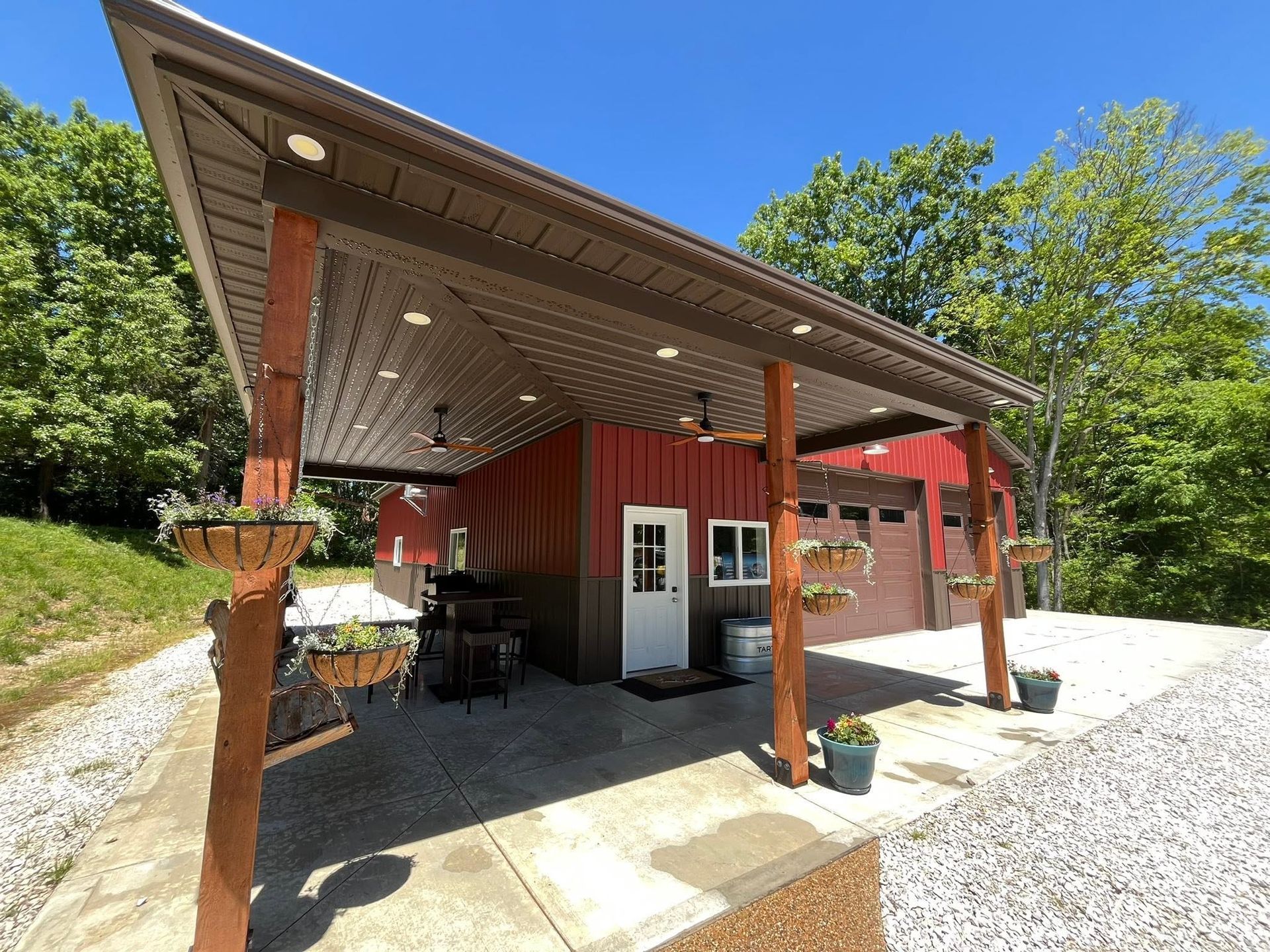 Red building with a covered porch and gravel driveway, surrounded by trees under a blue sky