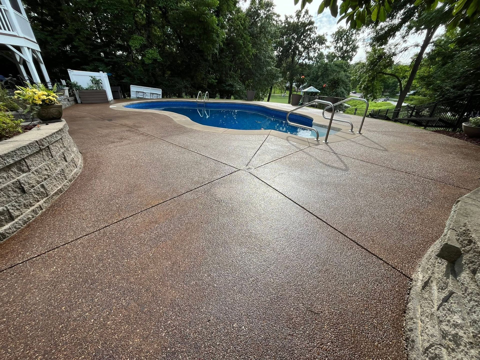 Blue swimming pool beside wet textured patio in a leafy outdoor area