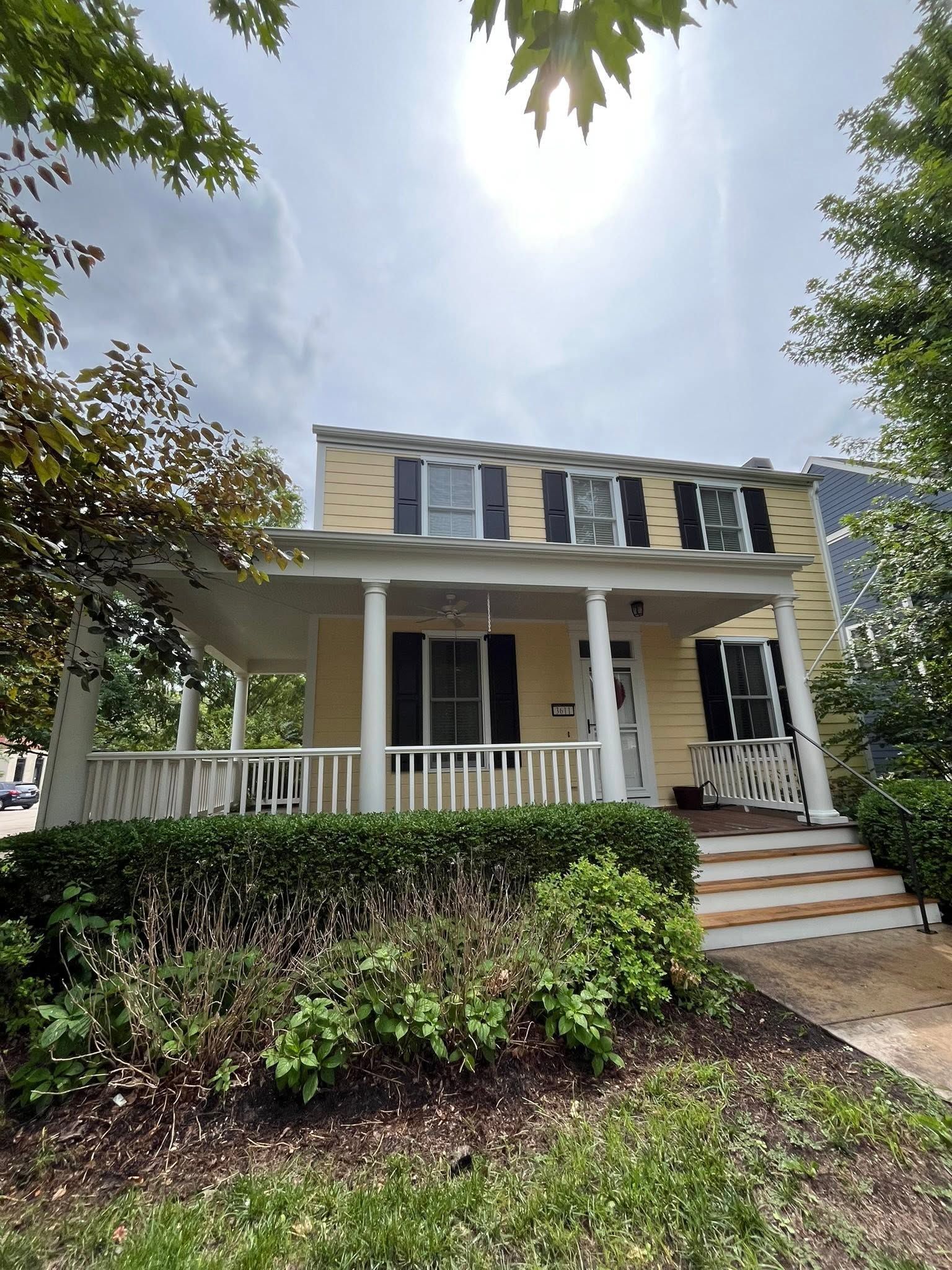 Two-story yellow house with white porch railing, black shutters, and brick steps surrounded by greenery