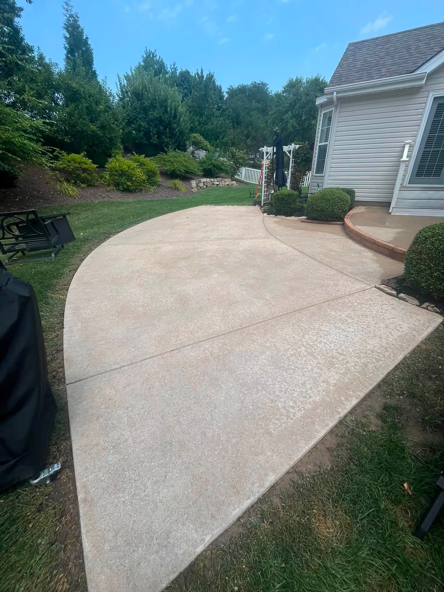 Curved concrete patio beside a white house, bordered by greenery and a grill cover.