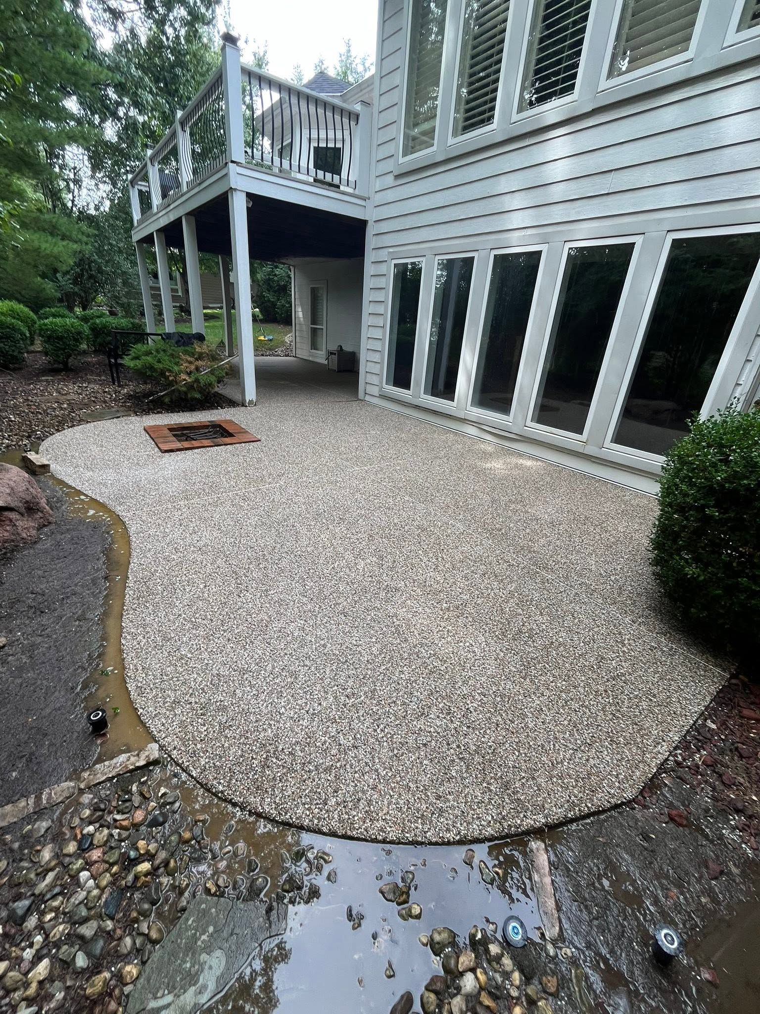 Gravel patio beside a gray house with large windows and a deck, bordered by shrubs and wet pavement.