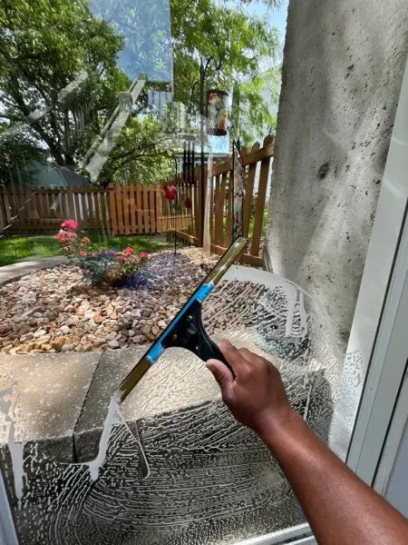 Person using a window squeegee to clean a large outdoor window with a backyard view.