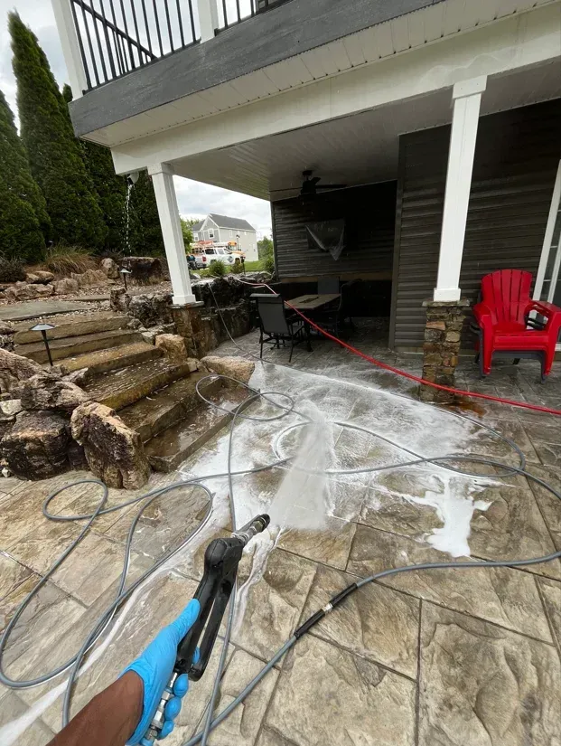 Power washing a stone patio beside a house, with water spraying across the ground and red chairs nearby