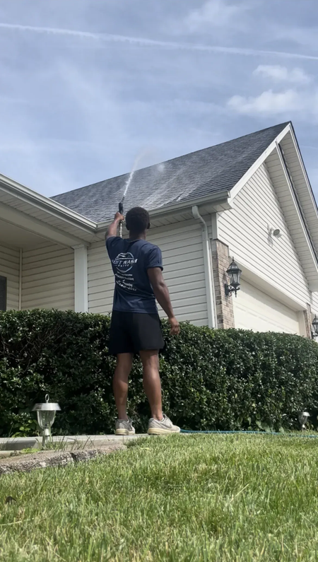 Person standing on a lawn reaching up toward a house roof with a long pole under a blue sky.