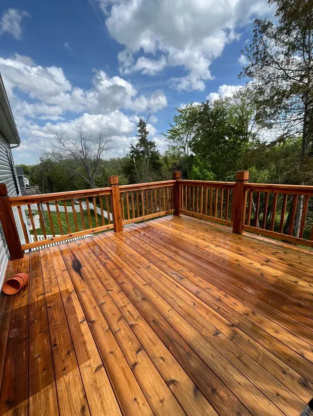 Freshly stained wooden deck with red railing under a partly cloudy sky, overlooking trees and a grassy yard