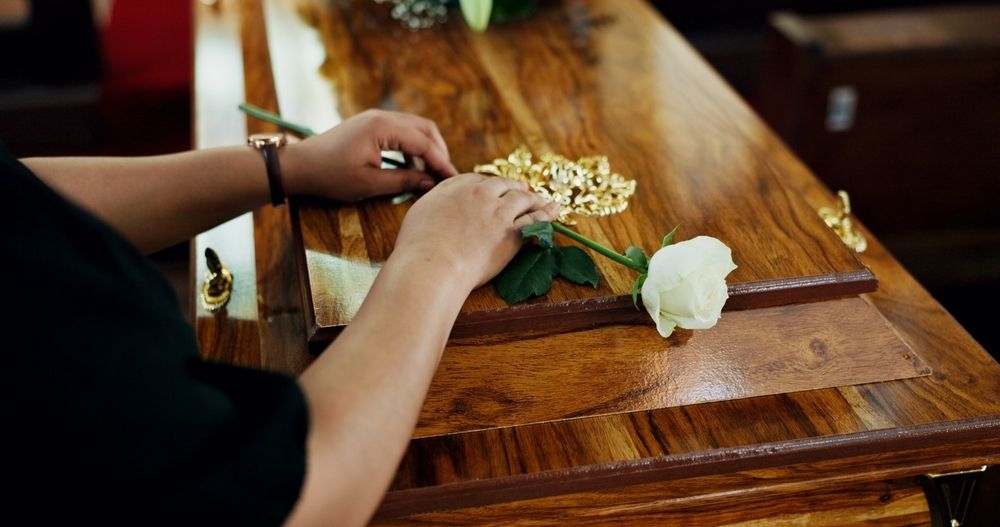 Hands placing a white rose on a wooden casket in a dim, indoor setting.