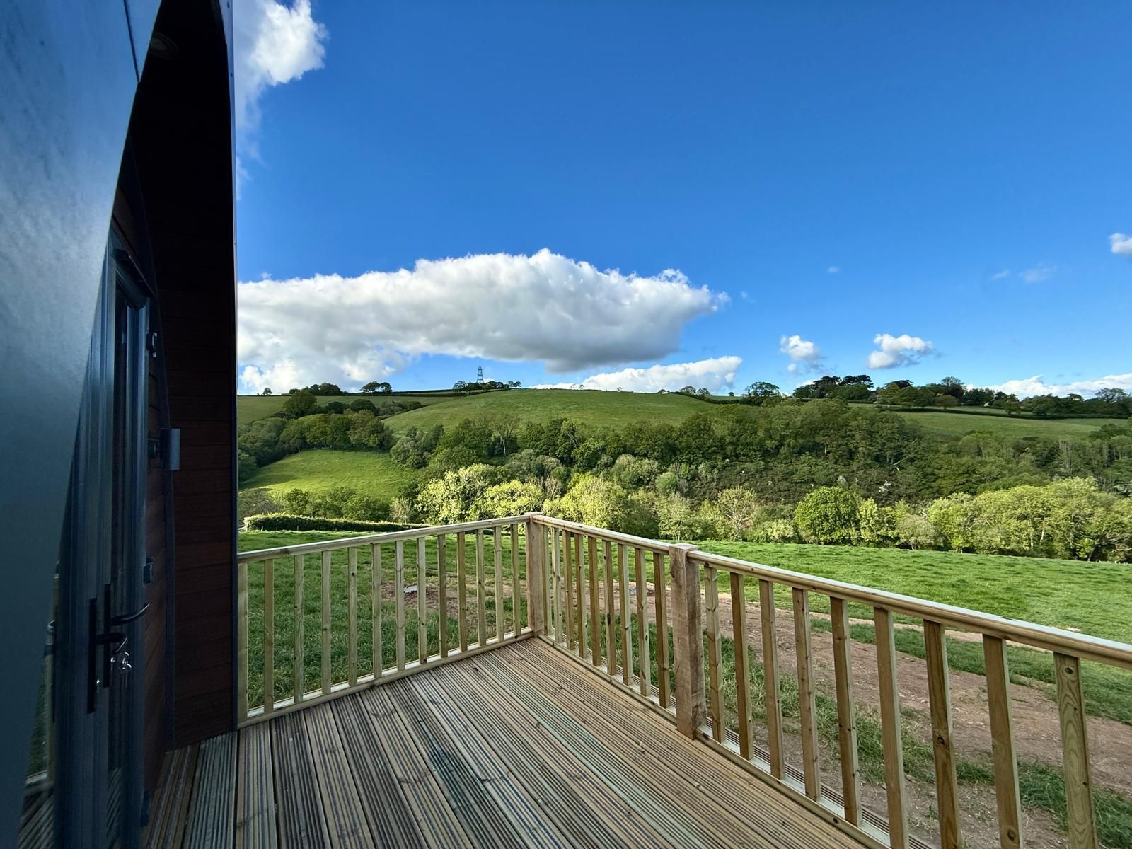 Wooden deck overlooking a lush green valley under a bright blue sky with clouds