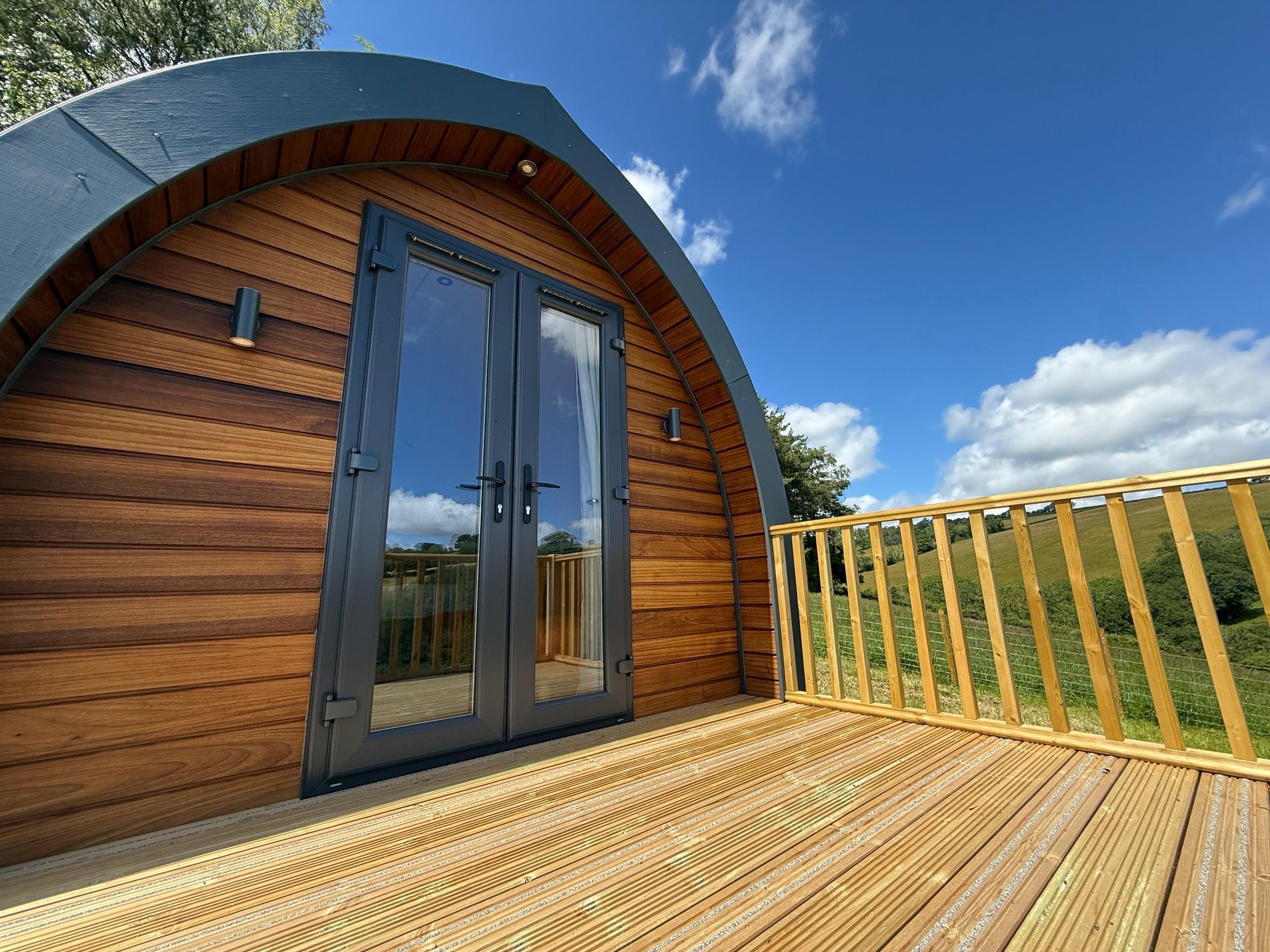 Small wooden cabin with arched roof and glass door on a sunlit deck under a blue sky