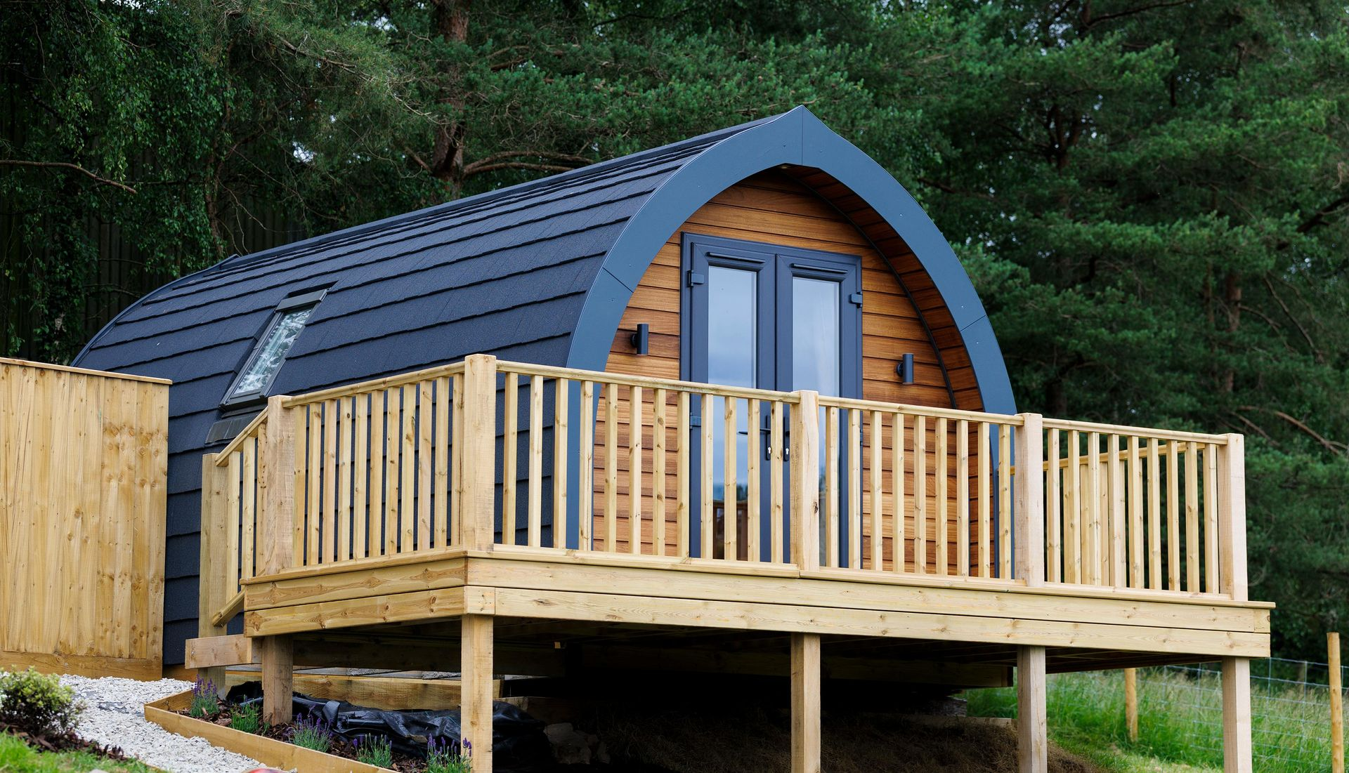 Small wooden cabin with a curved roof and front deck in a wooded setting