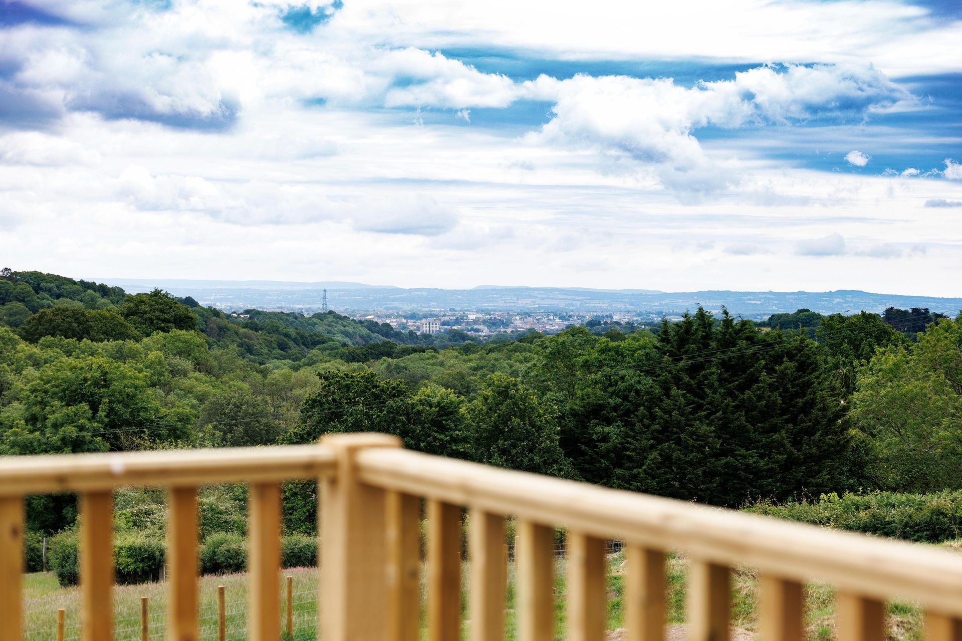 Wooden deck overlooking lush green hills and a distant city under a bright cloudy sky