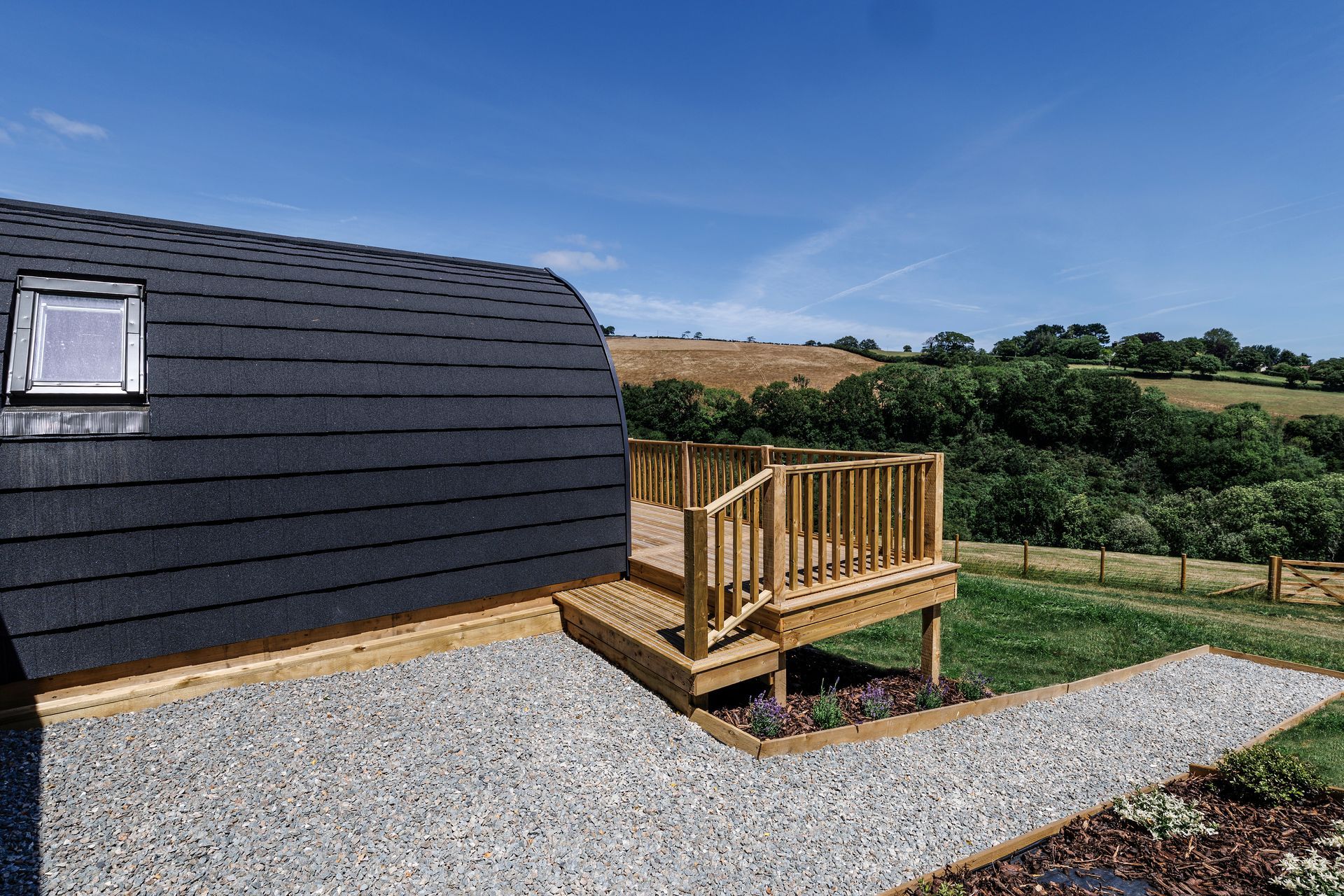 Gravel path beside a dark-roofed cabin leading to a small wooden deck overlooking green countryside