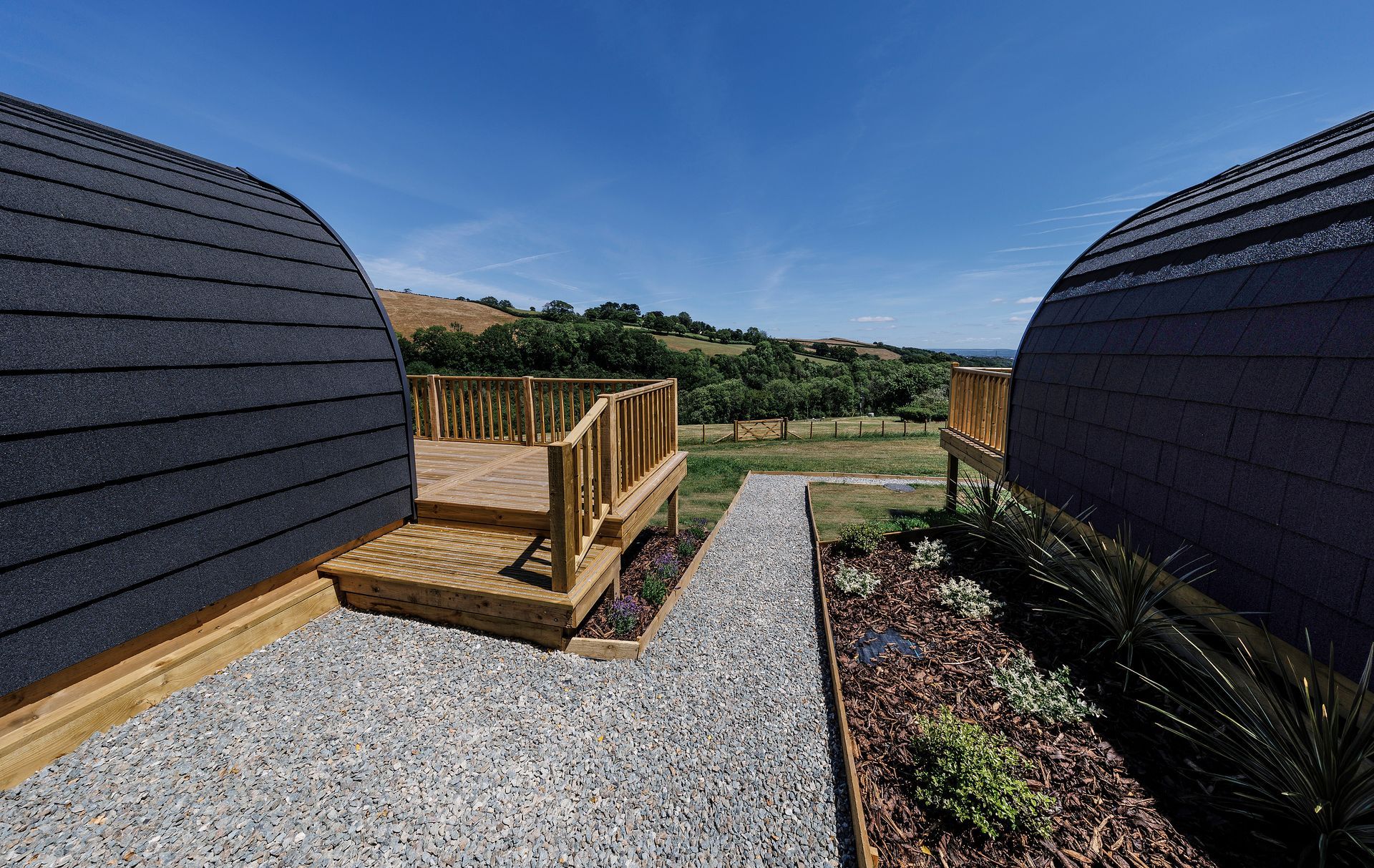 Gravel path between black dome cabins, with a small wooden deck overlooking a grassy field and blue sky.