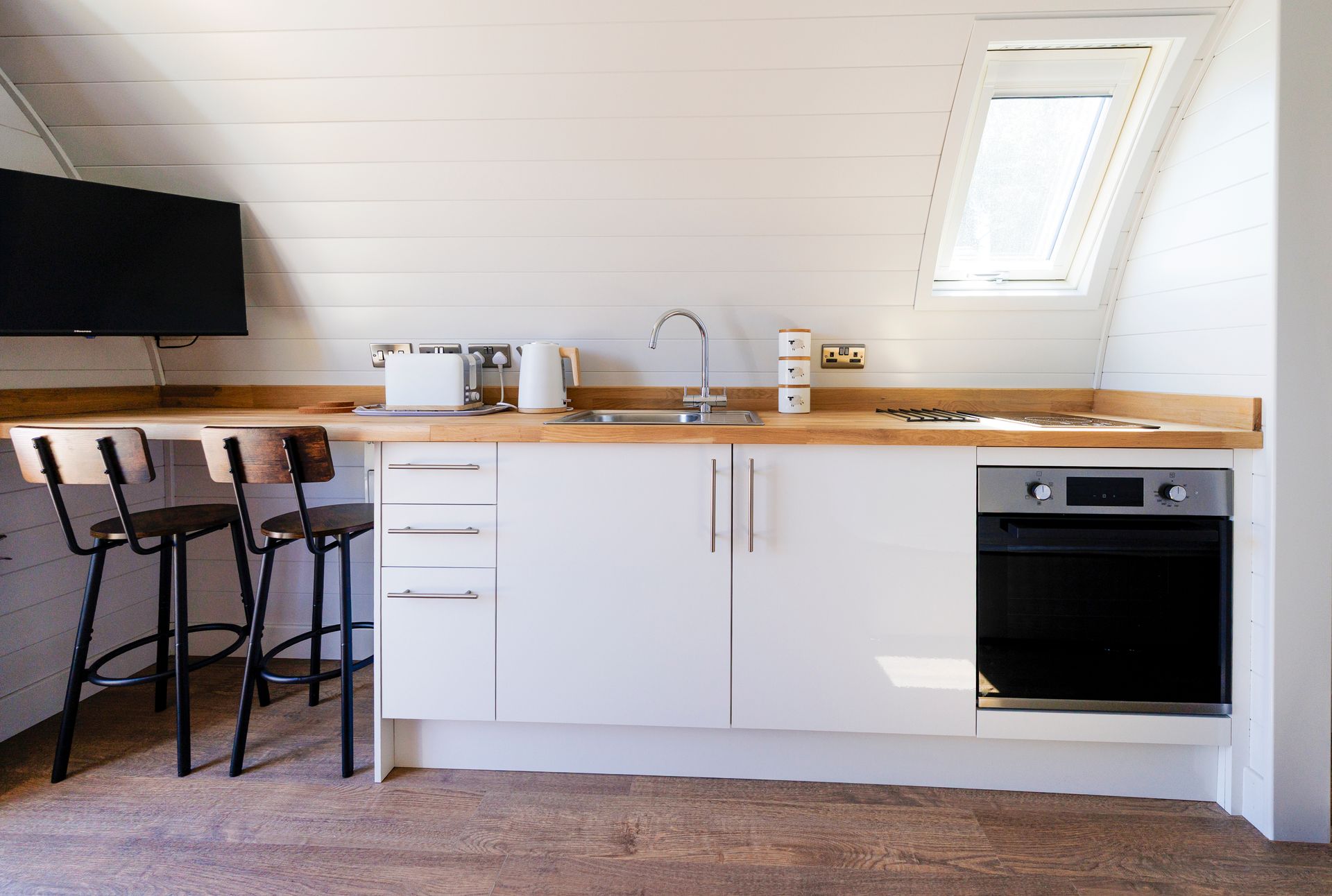 Modern white kitchen with wooden countertop, bar stools, sink, and built-in oven under a skylight