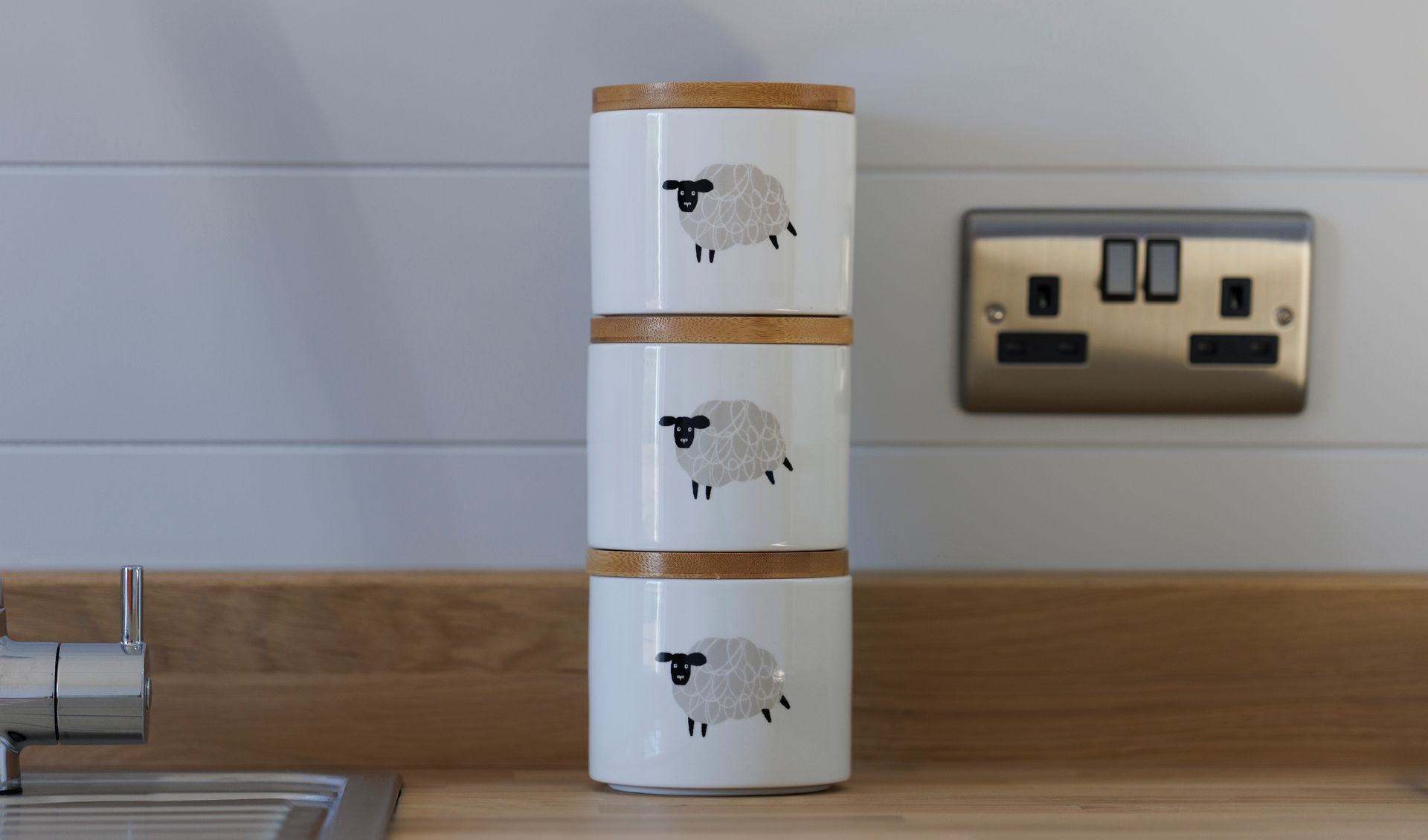 Three stacked white canisters with sheep illustrations on a wooden countertop beside a wall outlet.