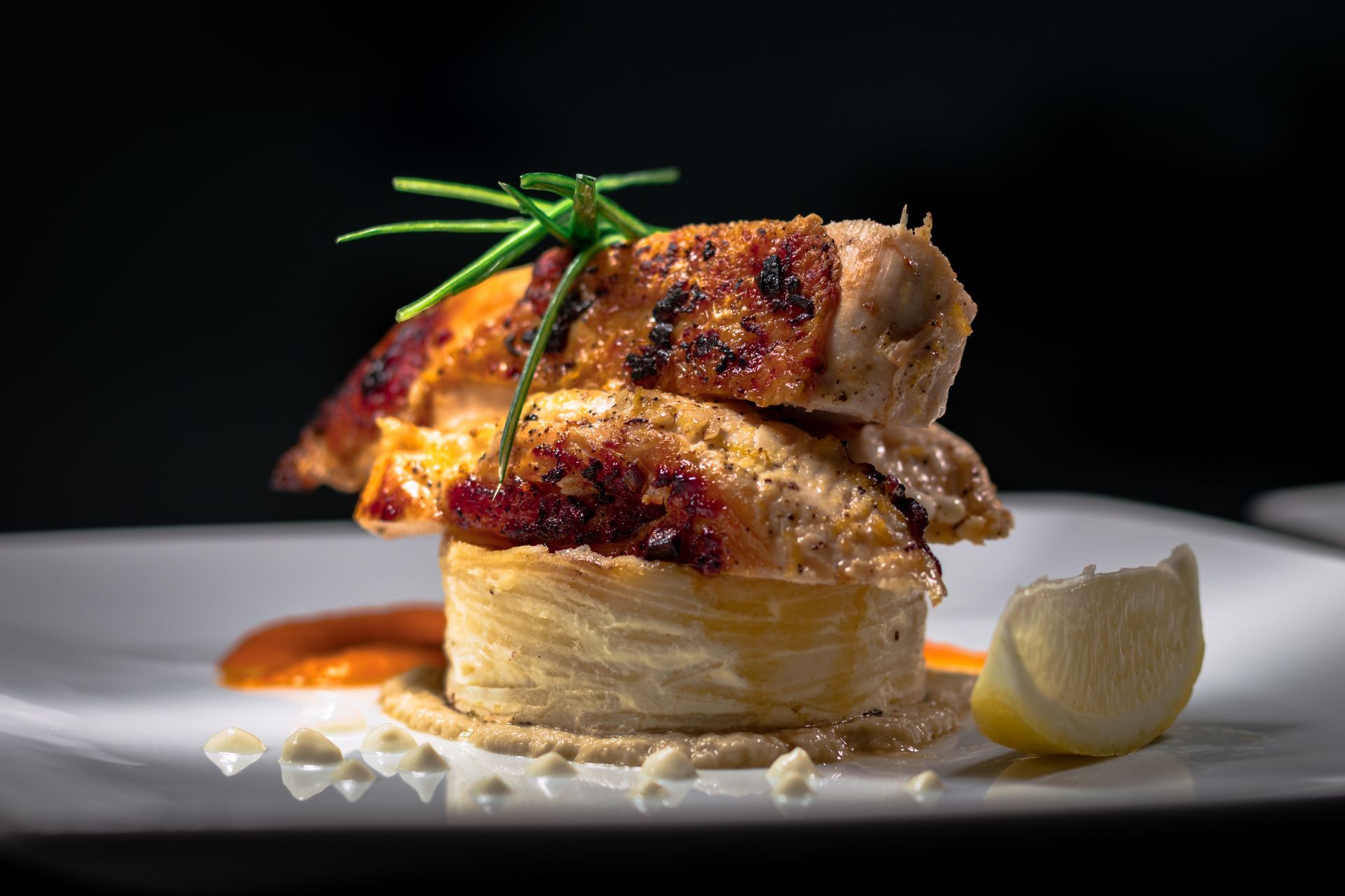 Seared stacked meat dish on a white plate, garnished with herbs and lemon, against a dark background