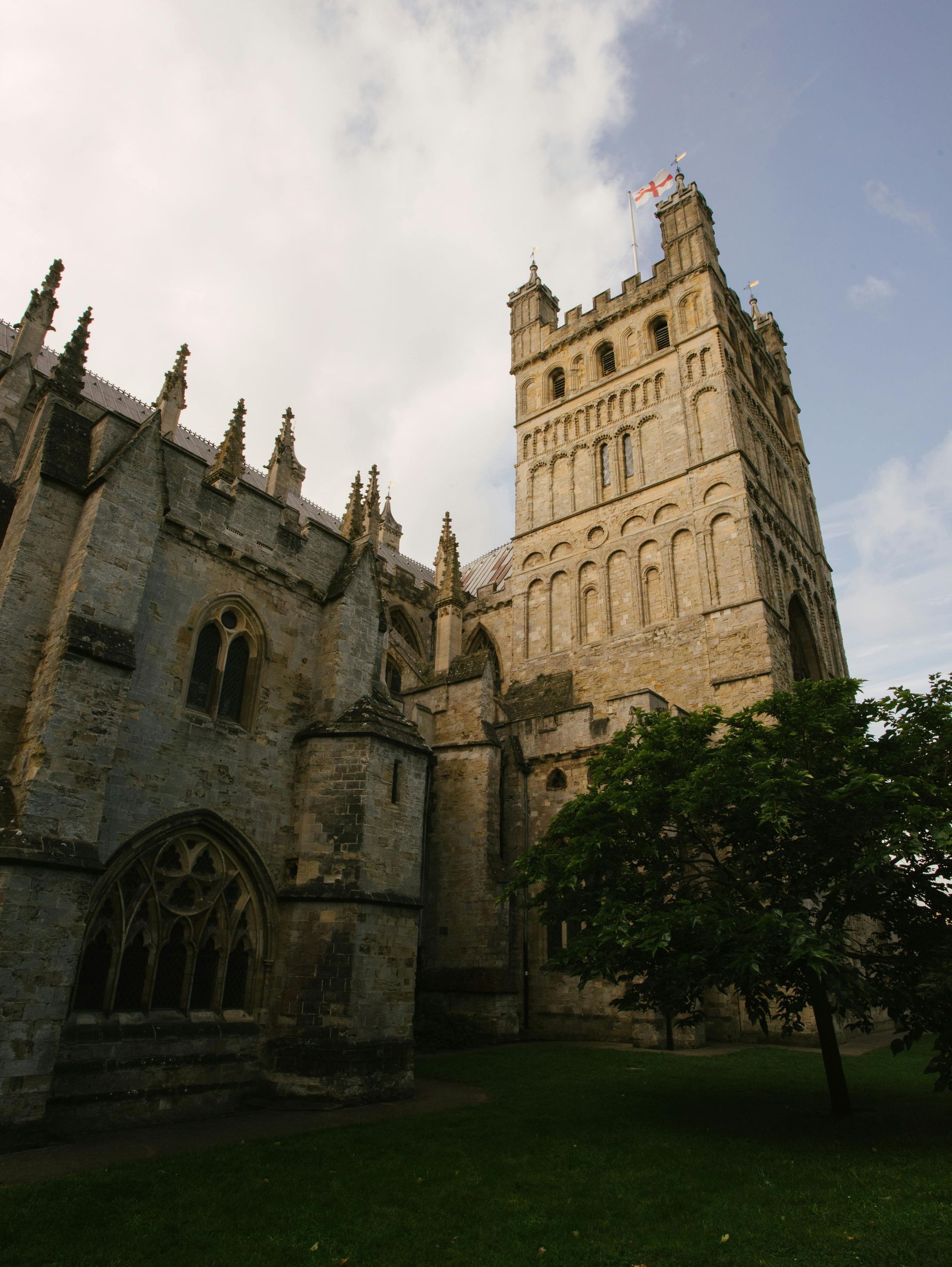 Stone cathedral tower and arched walls beside a grassy courtyard under a cloudy sky