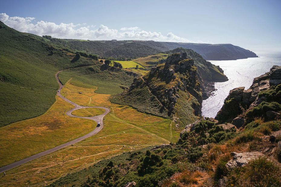 Coastal green cliffs with a winding road overlooking a sunlit bay and distant headland