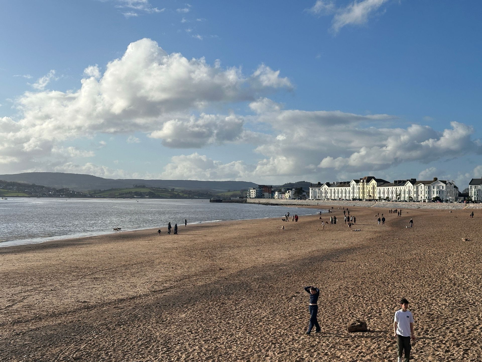 Wide sandy beach with people, calm sea, cloudy sky, and a coastal town in the distance