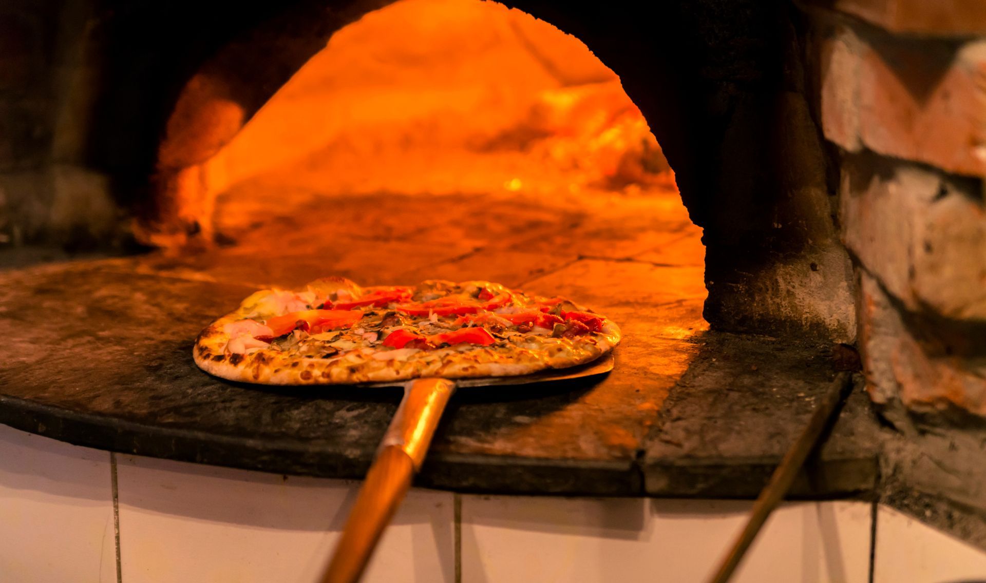 Pizza baking in a wood-fired oven on a peel, with orange flames glowing behind it
