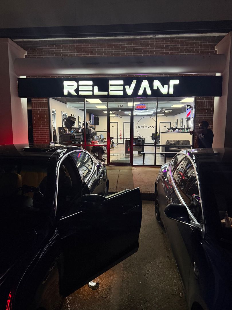 Relevant storefront at night, lit sign above glass entrance with cars parked in front