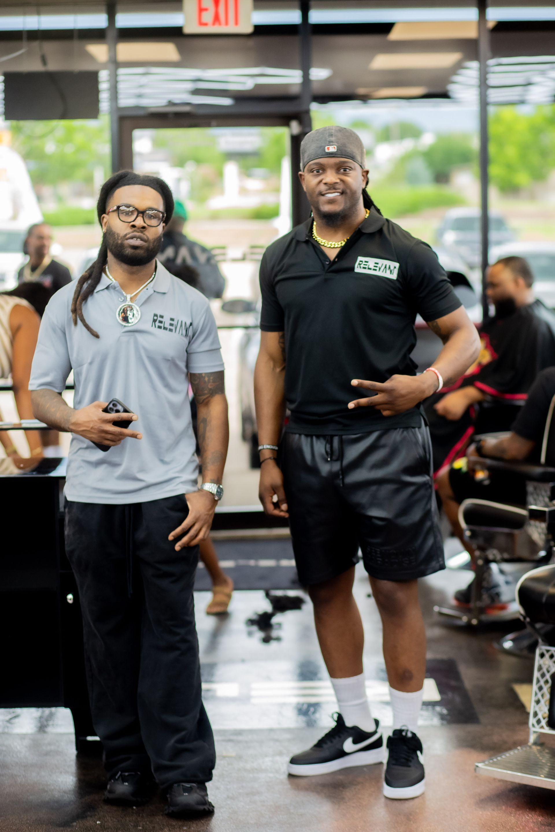 Two men standing in a waiting area, one holding a phone and one smiling in black athletic wear.