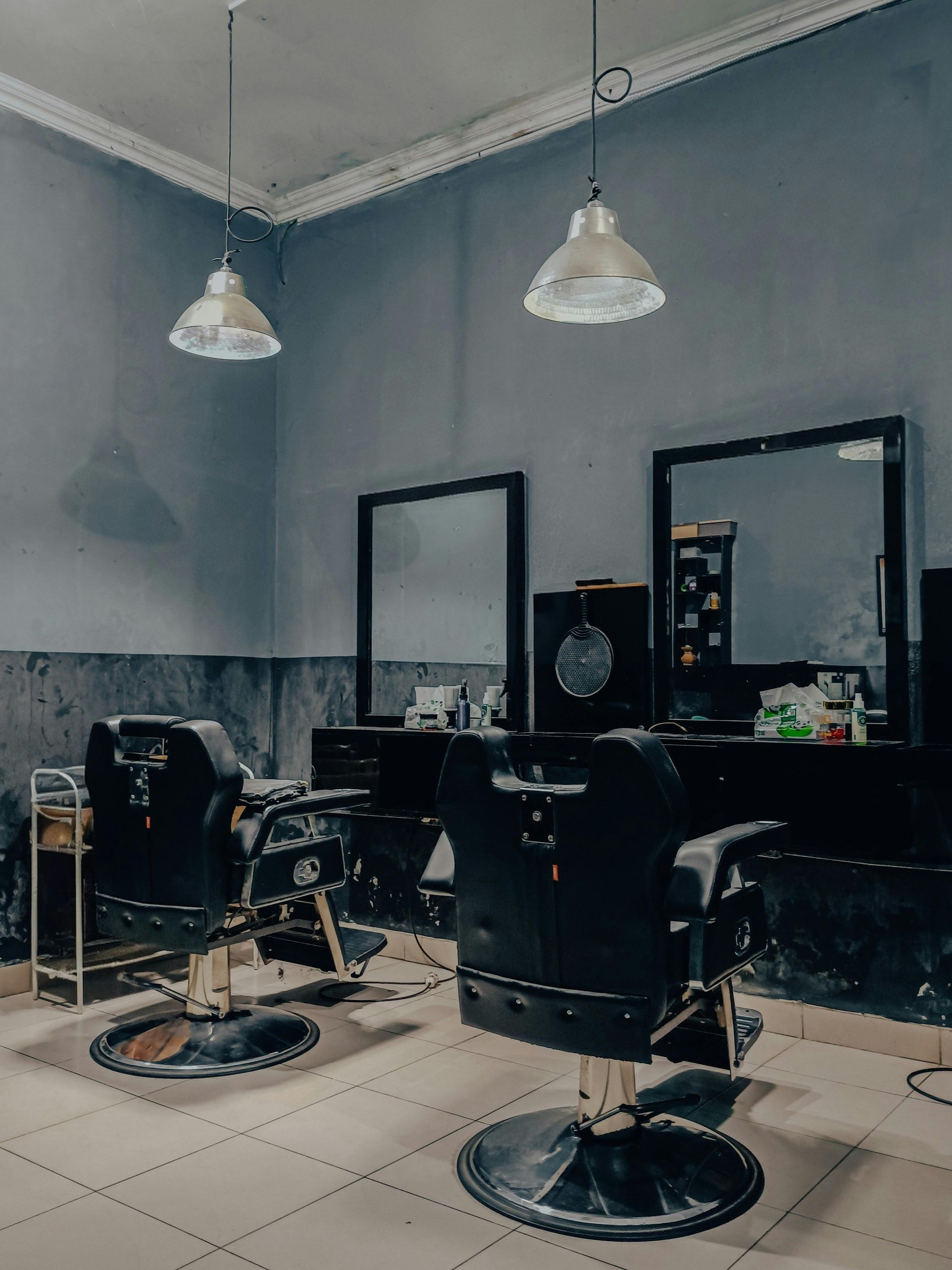 Empty barbershop with two black chairs, mirrors, and hanging pendant lights on tiled floor.
