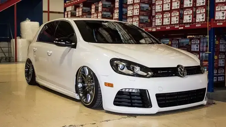 White Volkswagen Golf R with custom wheels, lowered suspension, in a warehouse setting.