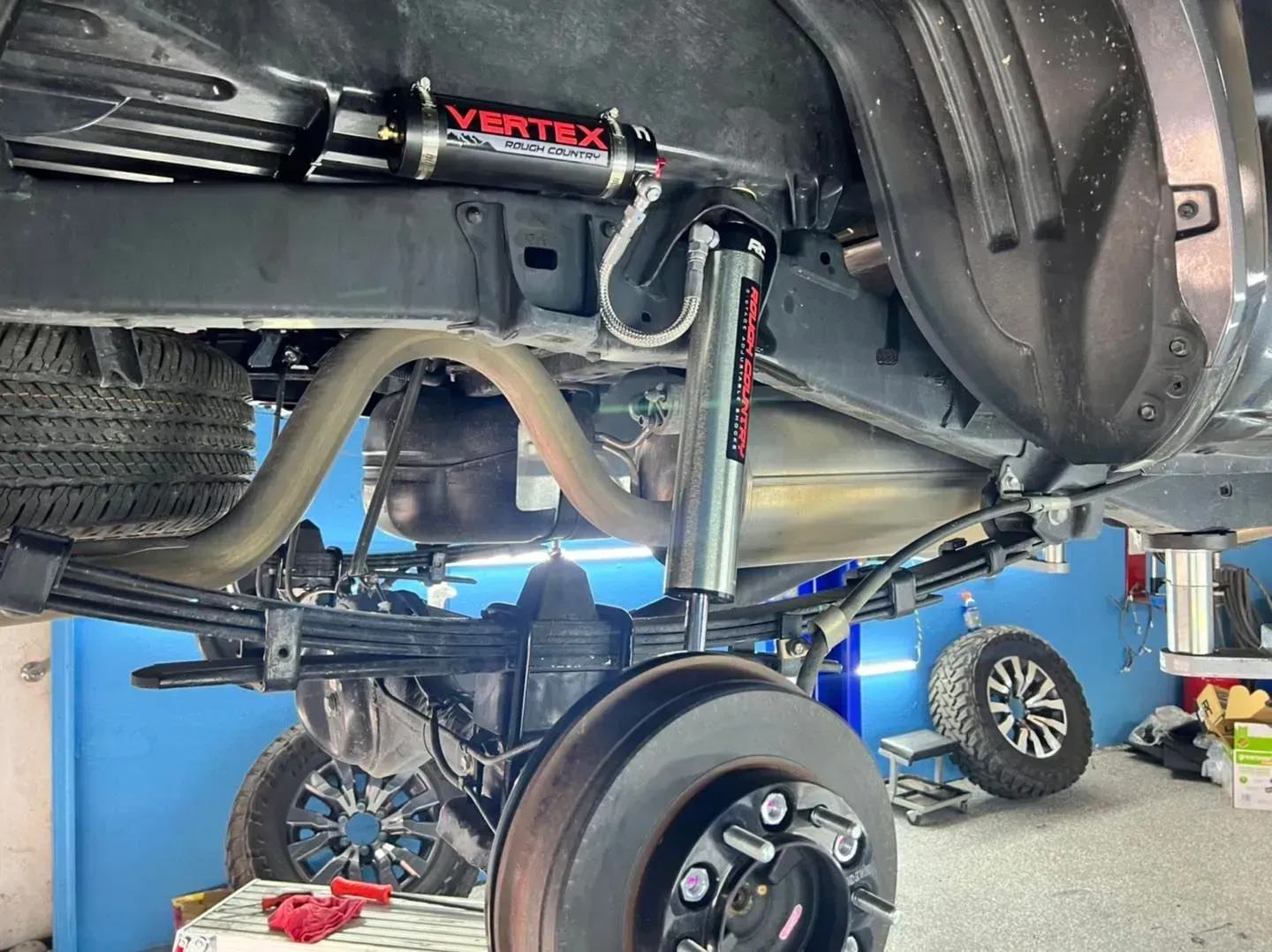 Underside view of a lifted truck rear suspension with Vertex shocks and custom modifications, in a garage setting.