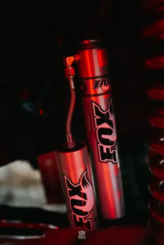Two Fox shocks, silver cylinders with red branding, connected by a hose, against a dark background.
