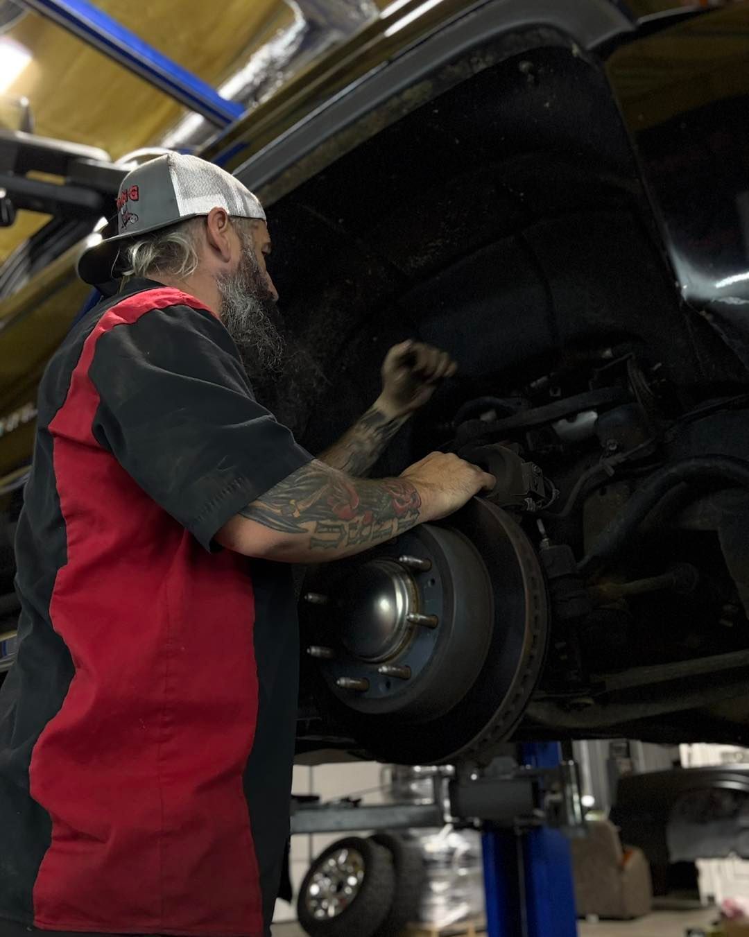 Mechanic working on a car's wheel, wearing a red and black shirt and hat, in an auto shop.