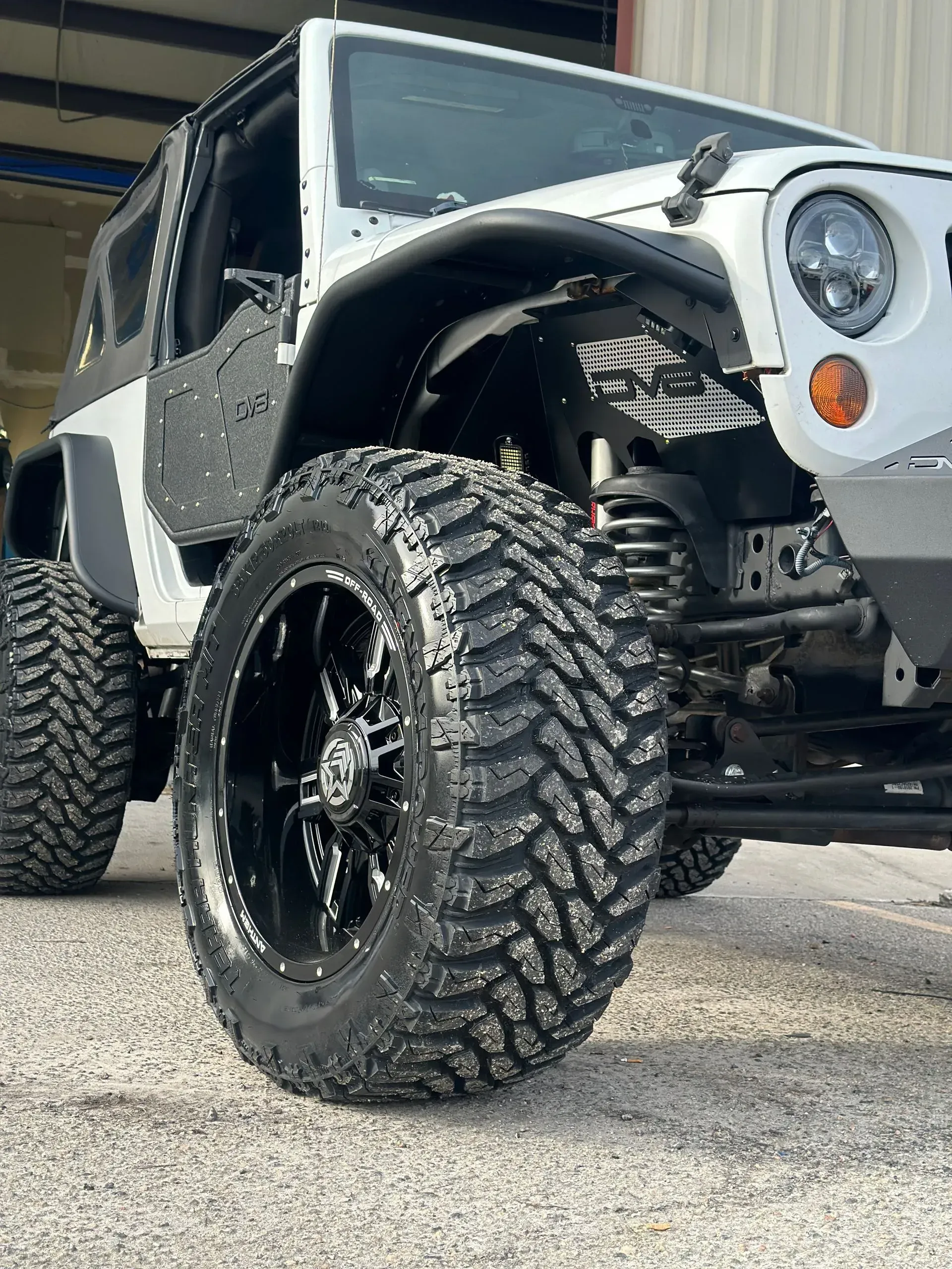 White Jeep Wrangler with large black tires, lifted, parked on gravel.