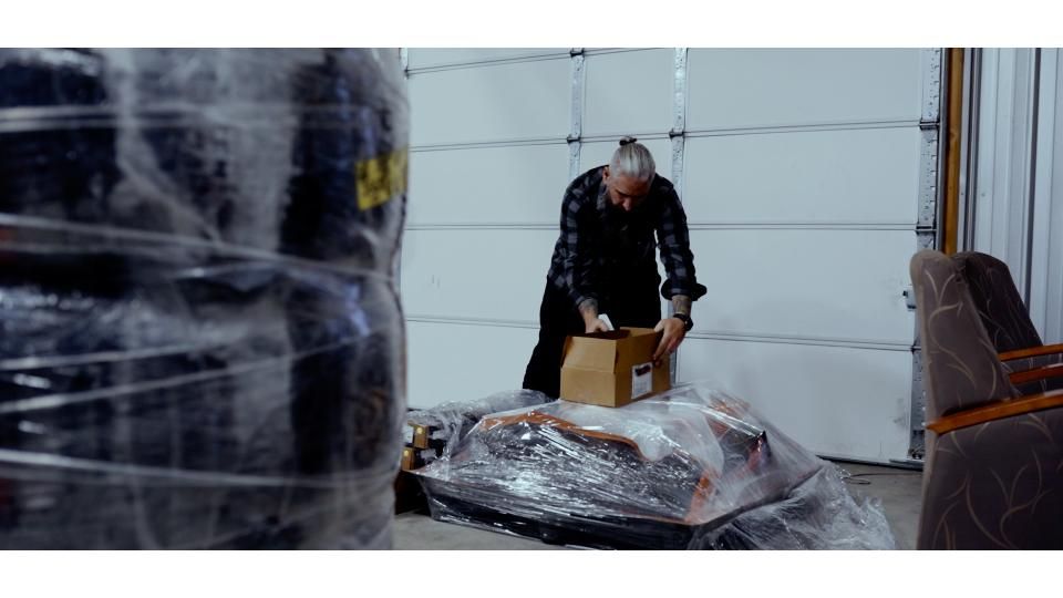 Man wrapping a large object in plastic in a garage, with stacked furniture and a wrapped item nearby
